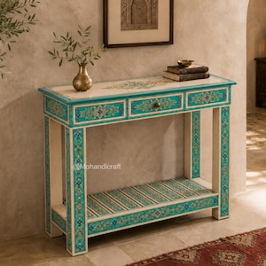 Peut inclure: Une console rectangulaire avec une finition crème et des motifs floraux complexes bleu sarcelle et dorés sur les façades des tiroirs, les pieds et l'étagère inférieure. La table comporte trois tiroirs avec de petits boutons ronds.