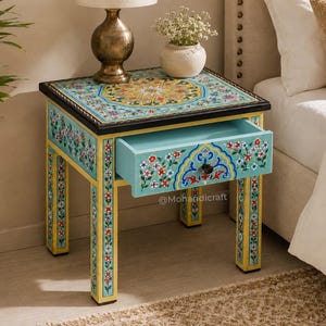 Può includere: Un tavolino quadrato in legno con un motivo floreale dipinto a mano nei toni del turchese, giallo e rosso. Il tavolo include un cassetto estraibile con un pomello nero e quattro gambe decorate con motivi floreali verticali e bordi gialli.