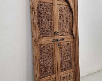 May include: A pair of intricately carved wooden doors with a traditional Moroccan design. The doors are made of light brown wood and feature a geometric pattern with a central arch.