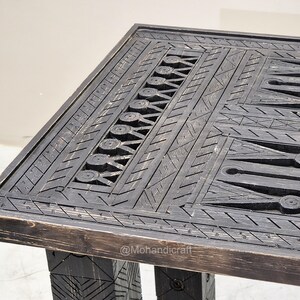 May include: A dark-stained wooden table with detailed carved geometric patterns. The rectangular tabletop features intricate designs, including circles, triangles, and linear elements. The table's legs are also carved, giving a rustic, handcrafted appearance.