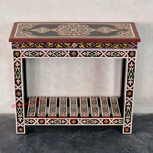 May include: A rectangular console table featuring a dark brown top with a central floral mandala design. The frame and lower slatted shelf are decorated with intricate geometric patterns in black, white, pink, and yellow paint throughout.