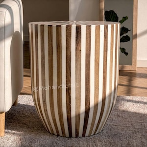 Puede incluir: Una mesa auxiliar cilíndrica con un patrón de rayas verticales que alterna madera marrón natural e incrustaciones blancas lisas. La superficie superior muestra un diseño radial de rayos de sol donde las rayas convergen en el centro.