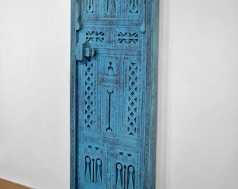 Op de afbeelding: Een turquoise, gesneden houten deurpaneel met ingewikkelde geometrische en gewelfde ontwerpen. Het paneel heeft een gedetailleerd slot en handvat en is tegen een witte muur gemonteerd. De algehele esthetiek suggereert een handgemaakt, decoratief item.