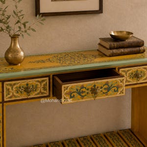 May include: A rectangular wooden console table with a yellow surface featuring intricate green and brown floral patterns, a light green border, and three drawers, one of which is open. The table has a lower slatted shelf and brass hardware.