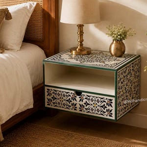 May include: A wall-mounted floating nightstand featuring a white finish with intricate black and gold floral inlay patterns on the drawer front and top surface, accented by dark green edges and a single open storage shelf with a circular cutout.