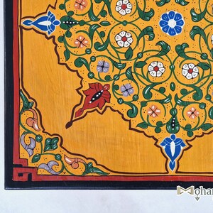 May include: Close-up of a hand-painted wooden surface with a vibrant floral design. The design features a yellow background with intricate patterns in green, red, white, blue, and purple. The border is black with a red accent. The corner of the surface is visible.