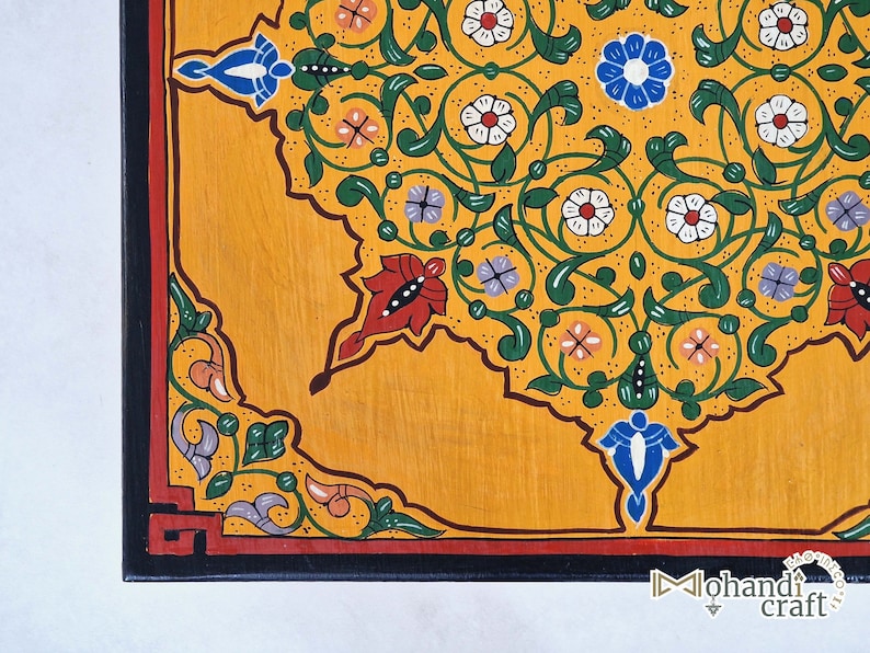 May include: Close-up of a hand-painted wooden surface with a vibrant floral design. The design features a yellow background with intricate patterns in green, red, white, blue, and purple. The border is black with a red accent. The corner of the surface is visible.