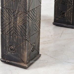 May include: Close-up of two dark wooden table legs. The legs are square and feature a geometric carved design. The wood has a distressed finish with visible grain and a dark, almost black colour. The legs are set on a light-coloured surface.