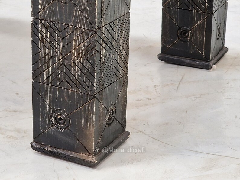 May include: Close-up of two dark wooden table legs. The legs are square and feature a geometric carved design. The wood has a distressed finish with visible grain and a dark, almost black colour. The legs are set on a light-coloured surface.