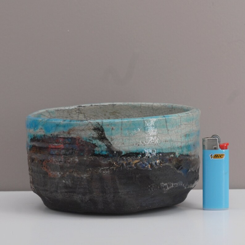 Puede incluir: Un cuenco grande de cer&aacute;mica ovalado con un esmalte craquelado. El cuenco tiene un esquema de color azul, blanco y negro con una superficie texturizada. El cuenco est&aacute; sobre una superficie blanca junto a un encendedor azul.