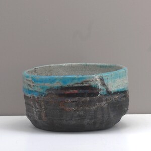 Puede incluir: Un peque&ntilde;o cuenco de cer&aacute;mica de forma ovalada con un exterior texturizado de color gris oscuro. El cuenco tiene un esmalte azul claro con un acabado craquelado y una fina banda de esmalte rojo.