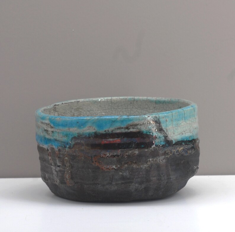 Puede incluir: Un peque&ntilde;o cuenco de cer&aacute;mica de forma ovalada con un exterior texturizado de color gris oscuro. El cuenco tiene un esmalte azul claro con un acabado craquelado y una fina banda de esmalte rojo.