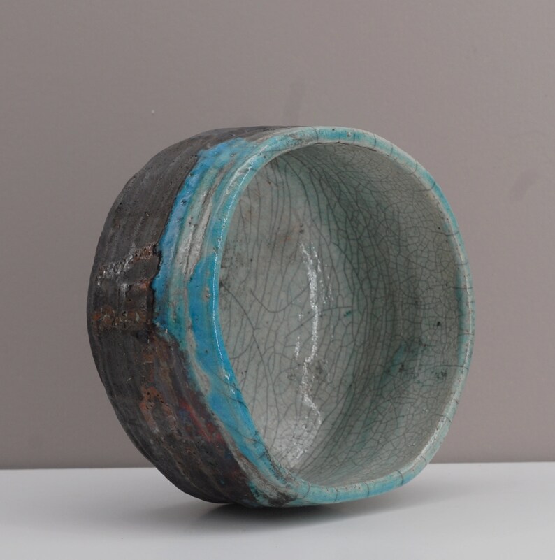 Puede incluir: Un cuenco de cer&aacute;mica con un esmalte craquelado en tonos de blanco y azul. El cuenco tiene un borde marr&oacute;n oscuro y una banda turquesa alrededor del borde.