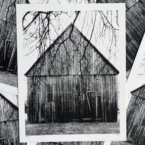 Könnte beinhalten: Schwarzweißfoto einer Holzscheune mit einem dreieckigen Dach. Die Scheune ist von kahlen Ästen umgeben.