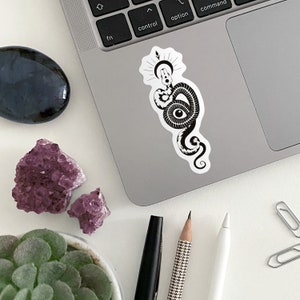 Op de afbeelding: Zwart-witte sticker van een slang met een oog in zijn lichaam, gewikkeld rond een halve maan met een ster erboven.