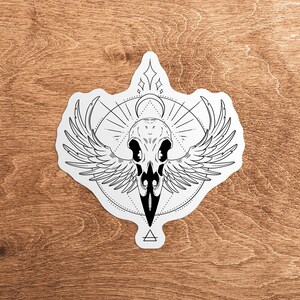 May include: Black and white illustration of a crow skull with wings spread wide. The skull is surrounded by a geometric design with a circle and a triangle.