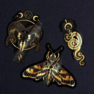 May include: Three gold foil stickers with black outlines. The first sticker features a ram skull with crescent moons and stars. The second sticker features a moth with a skull and moon phases. The third sticker features a snake with a crescent moon and stars.