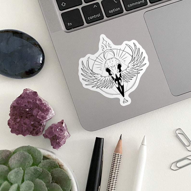 May include: Black and white sticker of a crow skull with wings and geometric designs on a silver laptop.