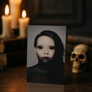 May include: A grayscale portrait of a person with dark hair, black eyes, and dark red lipstick. The image is on a card, with a skull and lit candles in the background, creating a spooky, gothic theme.