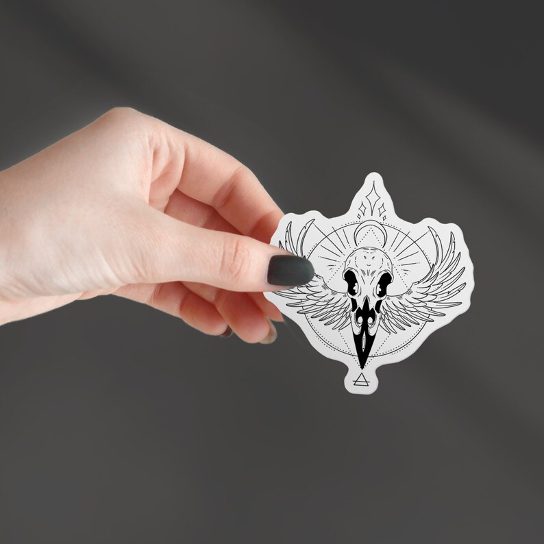 May include: Black and white sticker featuring a stylized crow skull with wings spread out. The skull is surrounded by a geometric design with a crescent moon and stars.