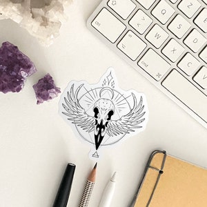 May include: A black and white sticker with a skull with wings and a geometric design. The sticker is on a white surface with a keyboard, pencils, and a notebook.