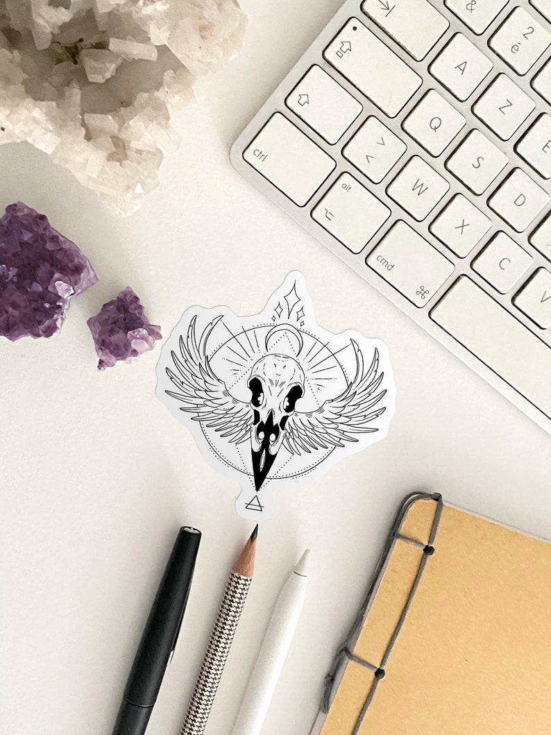 May include: A black and white sticker with a skull with wings and a geometric design. The sticker is on a white surface with a keyboard, pencils, and a notebook.