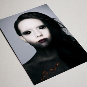 May include: A portrait of a person with pale skin, dark hair, and black eyes. The person's lips are a deep red, and the chest area has a fiery orange design. The background is a light gray.