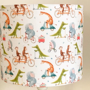 Puede incluir: Pantalla de lámpara blanca con un patrón caprichoso de animales montando bicicletas, patinetes y tándems. El diseño incluye jirafas, monos, caimanes y elefantes, con pequeños detalles florales. La pantalla es cilíndrica.