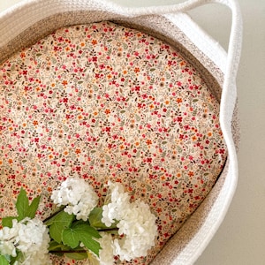 May include: A white woven basket with a handle, lined with a cream-colored fabric featuring a small floral pattern in shades of red, orange, and green. White flowers are placed inside the basket.