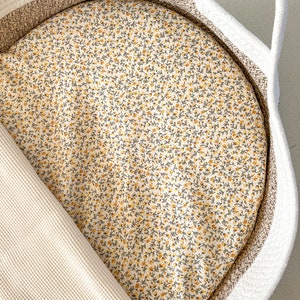 Peut inclure: Un panier en corde blanc avec un insert à motifs floraux. L'insert présente un fond crème avec de petites fleurs jaunes et des feuilles vertes. Une couverture en nid d'abeille crème est partiellement visible.