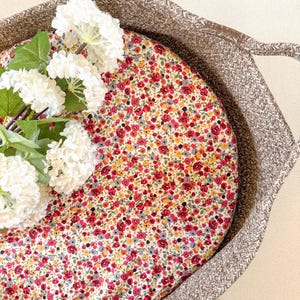 Peut inclure: Un panier en osier brun avec une doublure en tissu à motif floral blanc et rouge. Le panier a une poignée et est rempli de fleurs blanches.