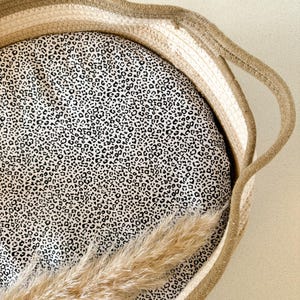 Peut inclure: Un panier rond en osier de couleur crème et marron. Le panier est doublé d'un tissu crème à imprimé léopard noir. Une longue plume d'herbe sèche brun clair est partiellement visible dans le panier.