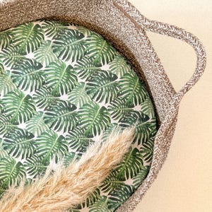Peut inclure: Un panier tissé brun avec une poignée, doublé d'un tissu blanc à motif de feuilles tropicales vertes. Une plume de pampa sèche repose à l'intérieur du panier.