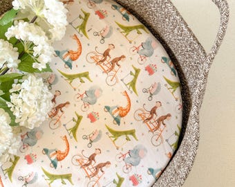 May include: A white fitted crib sheet with a repeating pattern of animals riding bicycles and scooters. The animals include a giraffe, an elephant, a monkey, and an alligator. The sheet is in a brown woven basket.