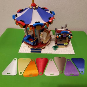 May include: A colorful Lego carousel with a blue and white canopy, red and white horses, and a small Lego stand with a green roof. The carousel is on a white base with a green surface.