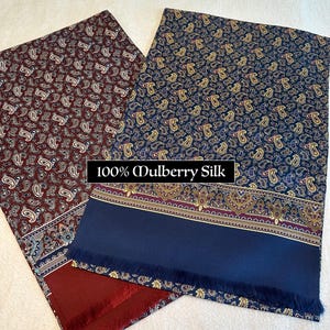 May include: Two silk scarves with paisley patterns. One is burgundy with white and gold accents, the other is navy blue with gold and purple accents. Both have a solid navy blue border with fringe. The text "100% Mulberry Silk" is visible.