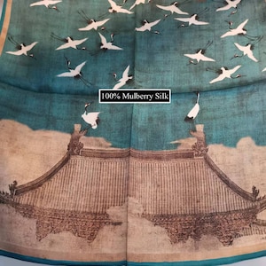 K&ouml;nnte beinhalten: Ein blauer Seidenschal mit einem wei&szlig;en und braunen Druck von Kranichen, die &uuml;ber einem traditionellen chinesischen Geb&auml;ude fliegen. Der Text "100% Mulberry Silk" ist auf dem Schal gedruckt.