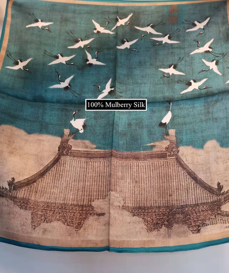 K&ouml;nnte beinhalten: Ein blauer Seidenschal mit einem wei&szlig;en und braunen Druck von Kranichen, die &uuml;ber einem traditionellen chinesischen Geb&auml;ude fliegen. Der Text "100% Mulberry Silk" ist auf dem Schal gedruckt.