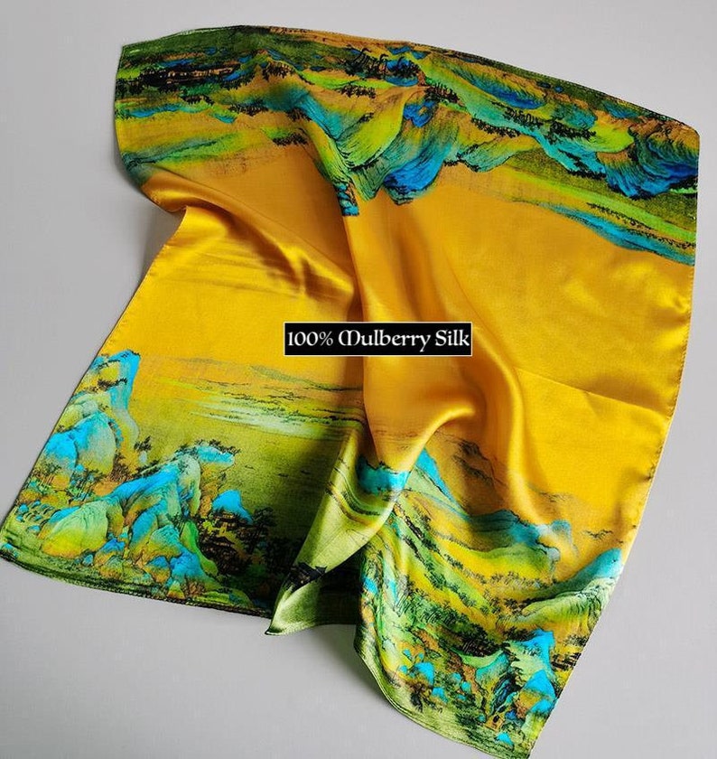 K&ouml;nnte beinhalten: Ein gelber Seidenschal mit einem Landschaftsdesign mit gr&uuml;nen und blauen Bergen und einem Fluss. Der Schal tr&auml;gt den Aufdruck "100% Maulbeersilk".