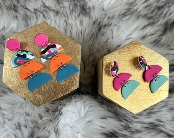 May include: A pair of colourful, abstract earrings. Each earring features a pink circle, a patterned half-circle, an orange half-circle, and a teal half-circle. The earrings are displayed on a gold hexagonal stand.
