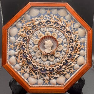 May include: Octagonal wooden frame with a blue background and a circular design of seashells. The center features a cameo of a woman's profile in a seashell.