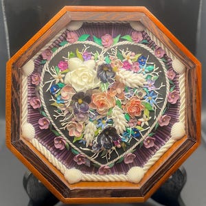 Könnte beinhalten: Ein gerahmtes herzförmiges Kunstwerk aus Muscheln, das einen Blumenstrauß mit einer weißen Rose in der Mitte darstellt. Der Rahmen ist achteckig und aus dunklem Holz.