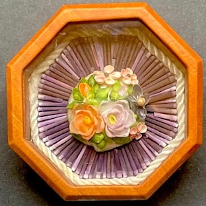 May include: A small wooden box with a glass top, displaying a floral arrangement made of seashells. The flowers are pink, white, and orange, and the background is made of purple and white seashells.