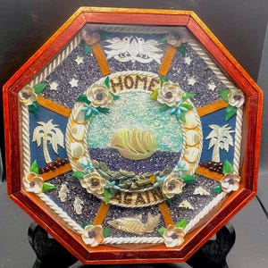 May include: Octagonal wooden frame with a red border and a rope design. The frame contains a mosaic of seashells, beads, and other materials depicting a ship sailing on the ocean. The mosaic also includes palm trees, flowers, and the words "HOME AGAIN".
