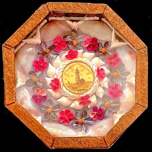 May include: A wooden octagonal box with a glass top, showcasing a seashell mosaic. The mosaic features a central image of a lighthouse, surrounded by pink and purple flowers made of seashells.