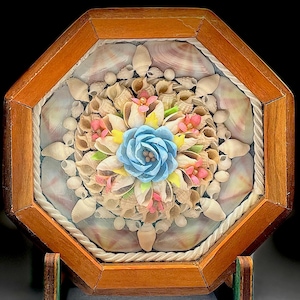 May include: An octagonal wooden framed seashell art piece. The center features a blue paper flower surrounded by seashells and smaller pink and yellow flowers. A rope-like border surrounds the seashell arrangement.