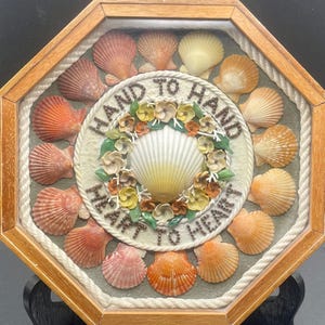 May include: A framed seashell art piece with a white seashell in the center surrounded by a wreath of colorful flowers. The frame is made of wood and has a rope border. The text "Hand to Hand Heart to Heart" is written in a circle around the seashell.