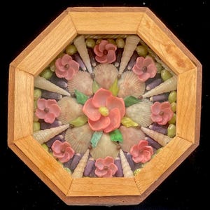 May include: A framed seashell mosaic with a circular design. The center features a large pink flower surrounded by smaller pink flowers and seashells. The frame is made of wood and has an octagonal shape.