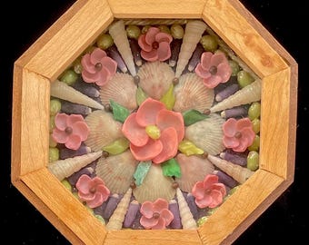 Könnte beinhalten: Ein gerahmtes Muschelmosaik mit einem kreisförmigen Design. Die Mitte zeigt eine große rosa Blume, umgeben von kleineren rosa Blumen und Muscheln. Der Rahmen ist aus Holz und hat eine achteckige Form.