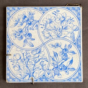 May include: Square ceramic tile with four circular designs featuring blue floral and fruit illustrations on a white background. The tile has a decorative border and a metal hanging loop.